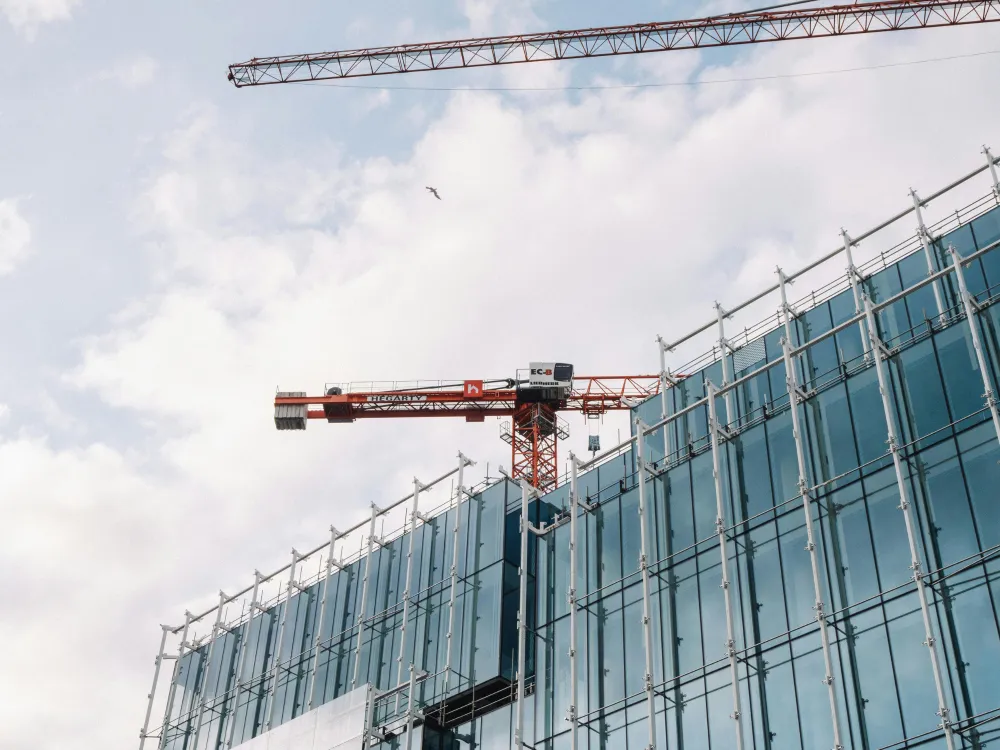 A construction site with a red crane atop a modern glass building, against a cloudy sky.