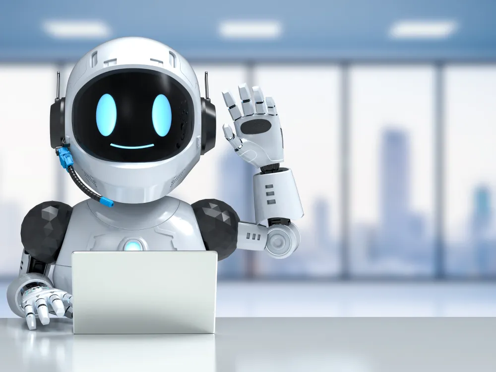 A friendly robot with a white and black design gestures hello while sitting at a desk with a laptop in a modern office setting.