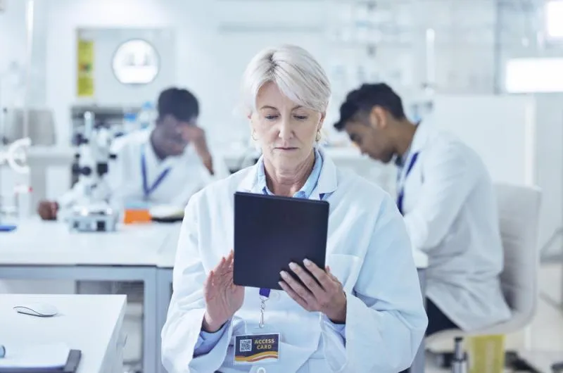 A scientist in a lab coat holds a tablet, surrounded by laboratory equipment and colleagues working in the background.