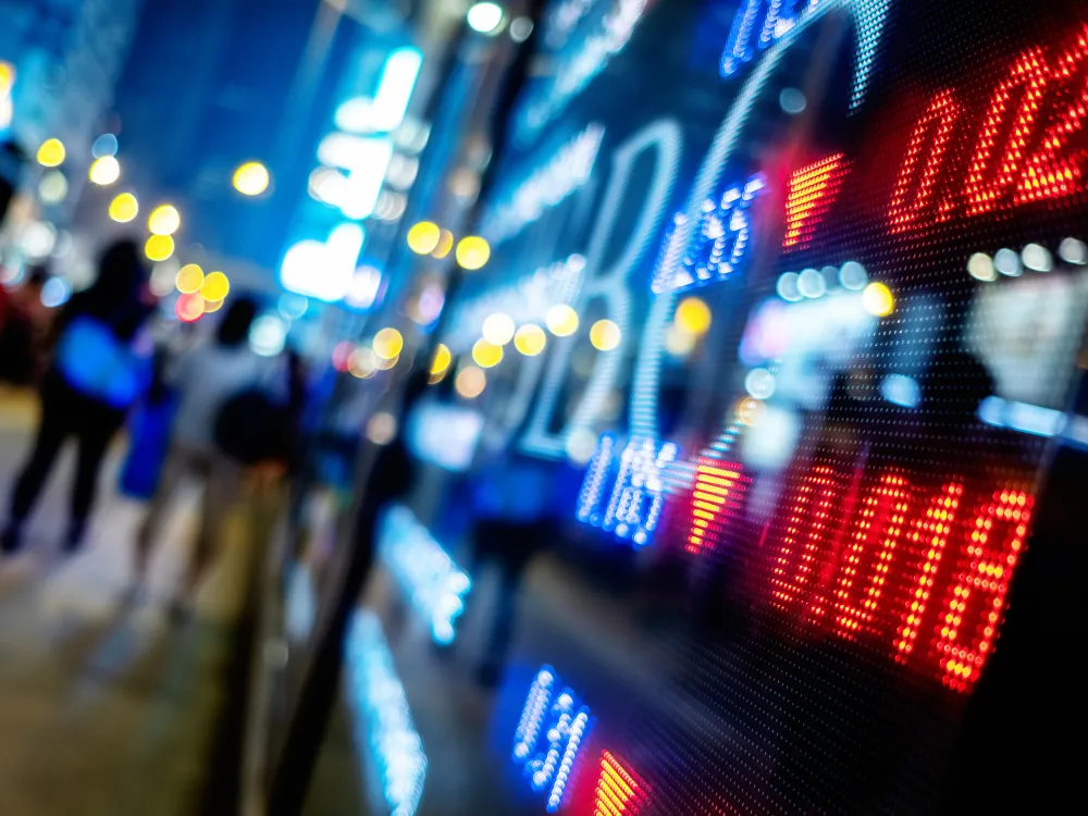 A digital stock market board displays red downward arrows and numbers at night with blurred city lights and pedestrians in the background.