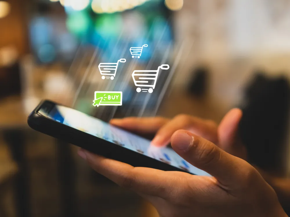 A person uses a smartphone, with digital shopping cart icons and a "Buy" button overlaid, representing online shopping or mobile e-commerce.