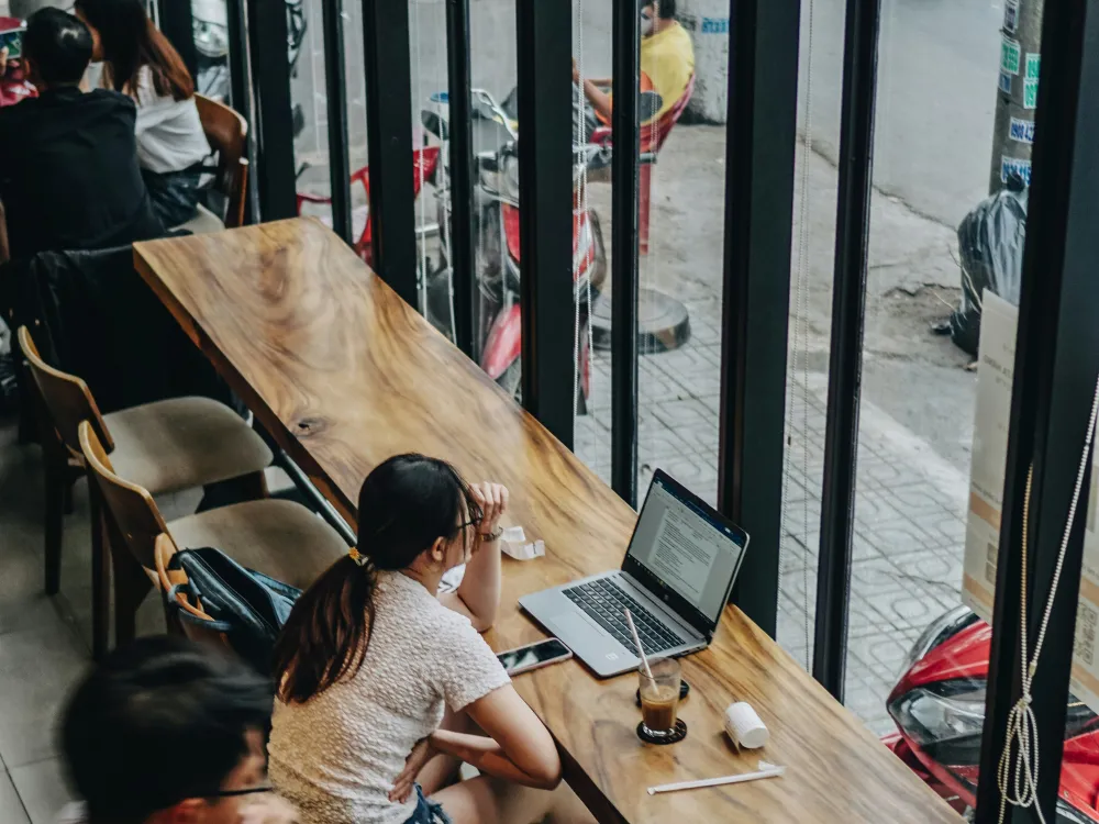 Woman working on a laptop in a café by a window, illustrating remote work and digital nomad lifestyle.