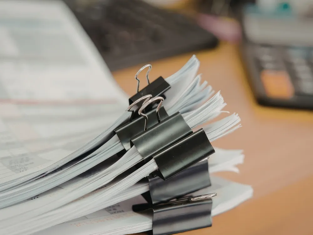 Stacks of documents secured with large black binder clips are arranged on a desk, with a blurred keyboard and calculator in the background.