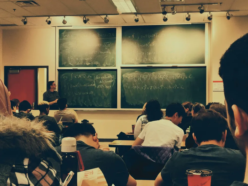 A college classroom during a math lecture, with a professor standing at the front writing equations on large chalkboards while students sit facing forward, taking notes.
