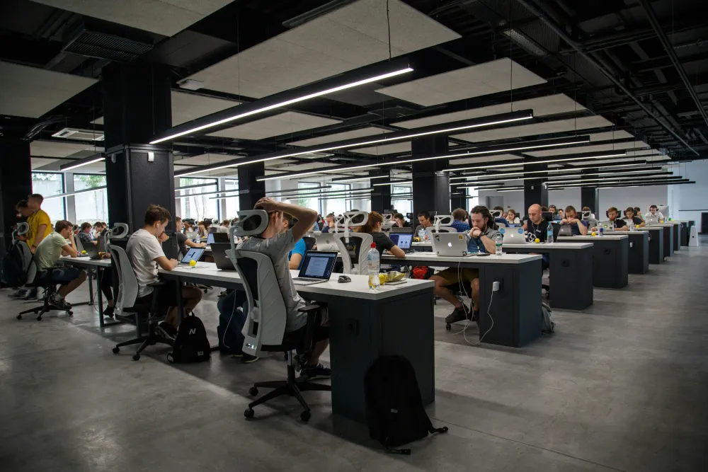 Modern open-plan office with rows of developers working on laptops at shared desks under sleek ceiling lighting.