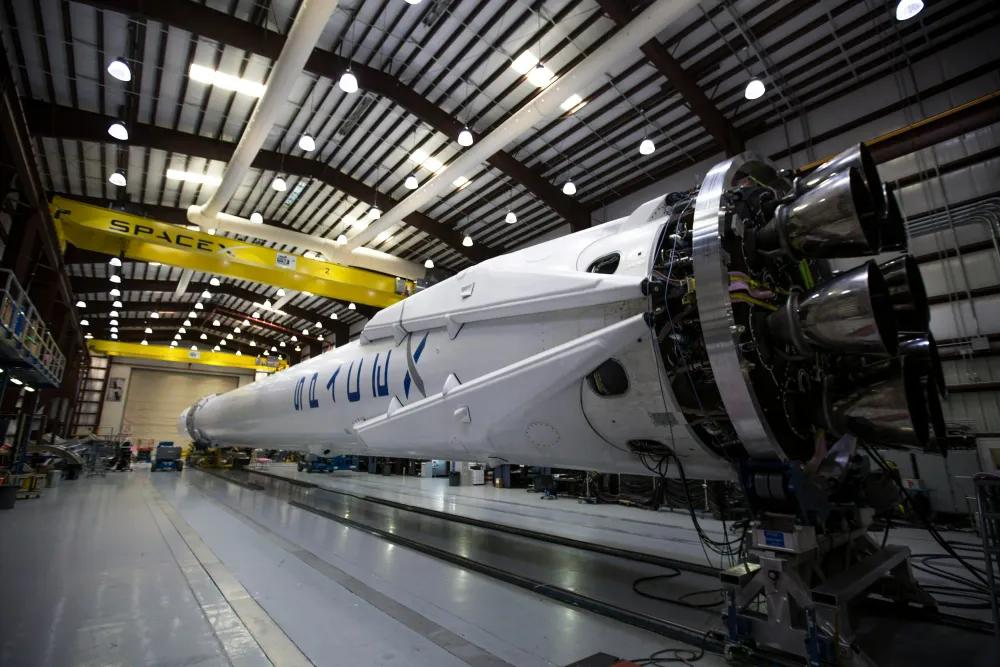 SpaceX rocket in a large assembly hangar, showing engines and fuselage under bright industrial lighting.