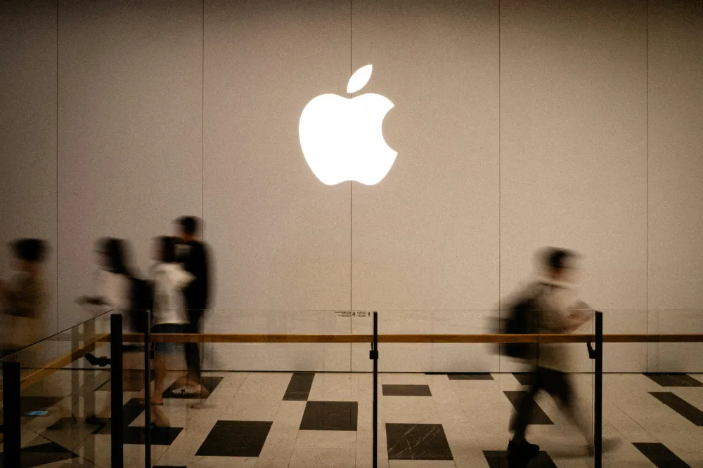 Blurred shoppers walk beneath a glowing Apple logo inside a modern Apple Store with a polished black-and-white tiled floor