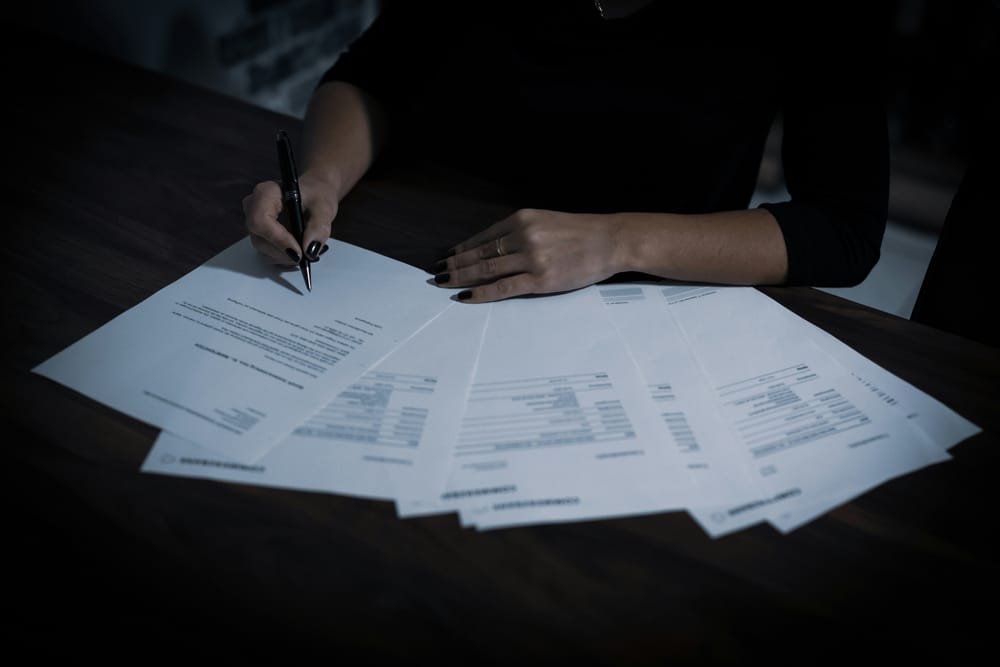A person's hands signing financial documents spread across a dark desk