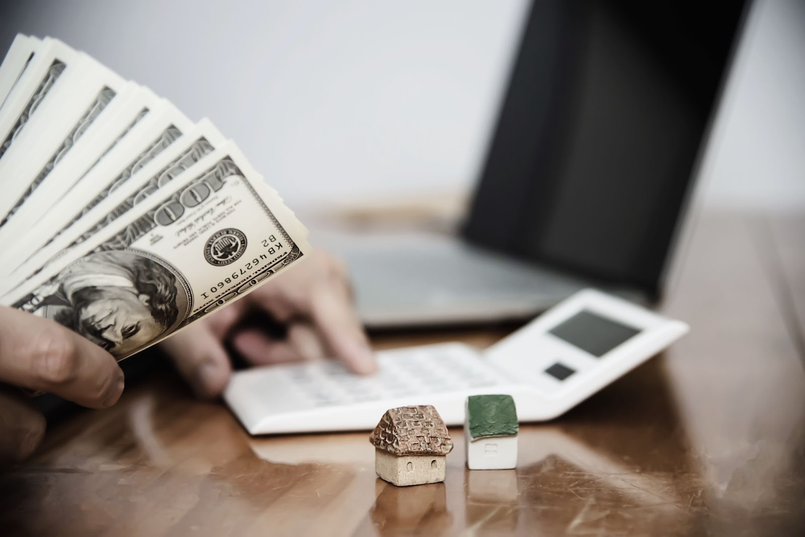 A person holding a fan of hundred dollar notes while using a calculator, with two small model houses on the desk