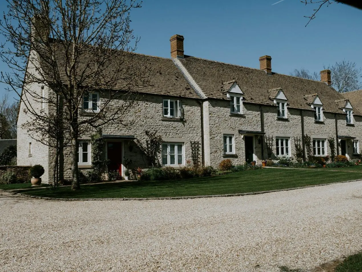 A row of charming stone cottages with manicured lawns and flower beds, set against a clear blue sky. The gravel driveway leads to the houses.