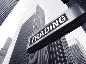 A close-up view of a sign reading "TRADING" against a backdrop of tall, modern skyscrapers in a black and white setting.