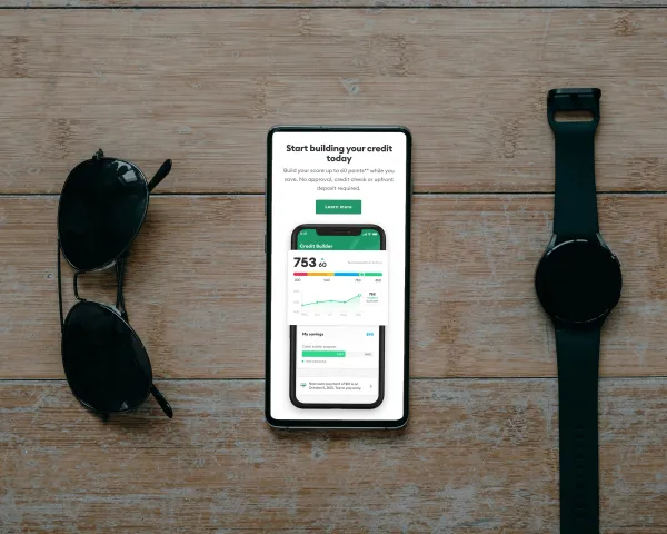 A smartphone displays a credit-building app, flanked by sunglasses and a smartwatch on a wooden surface.