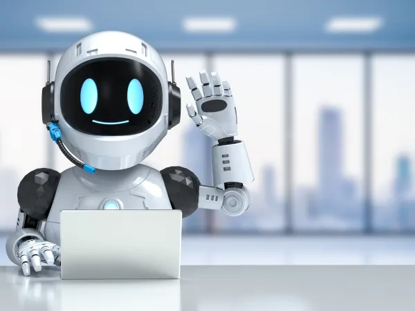 A friendly robot with a white and black design gestures hello while sitting at a desk with a laptop in a modern office setting.