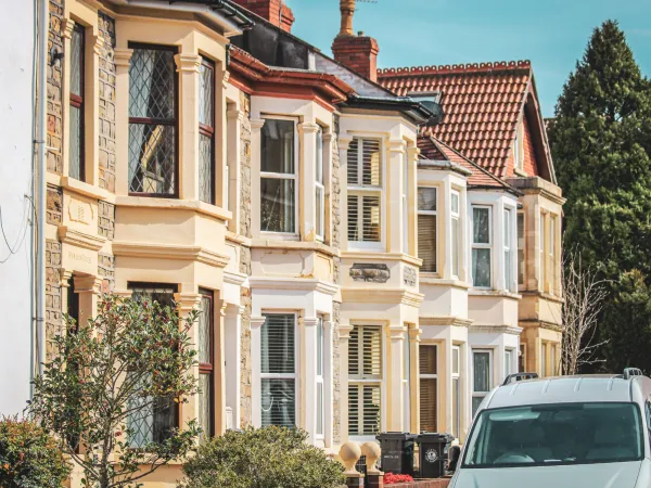 Row of charming Victorian-style houses with large bay windows, a white van parked in front, and greenery in the foreground.