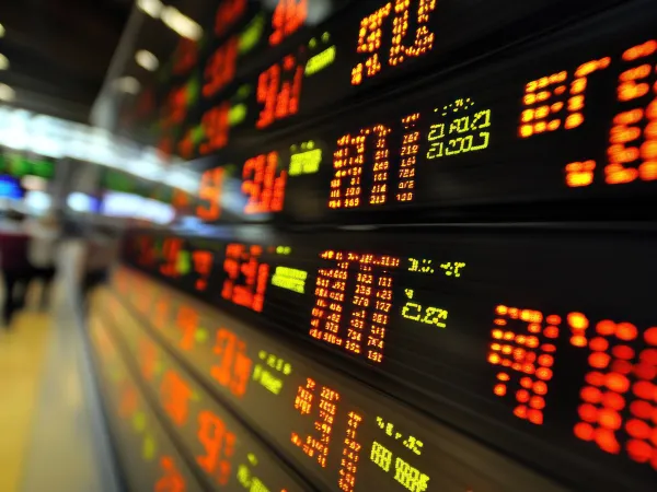 A blurred stock market display shows fluctuating prices in bright red and green digits, with traders visible in the background.