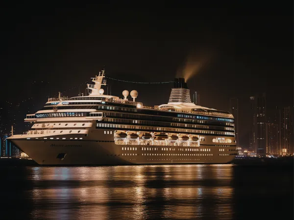 A large cruise ship illuminated at night, docked peacefully with city lights reflecting on the water's surface.