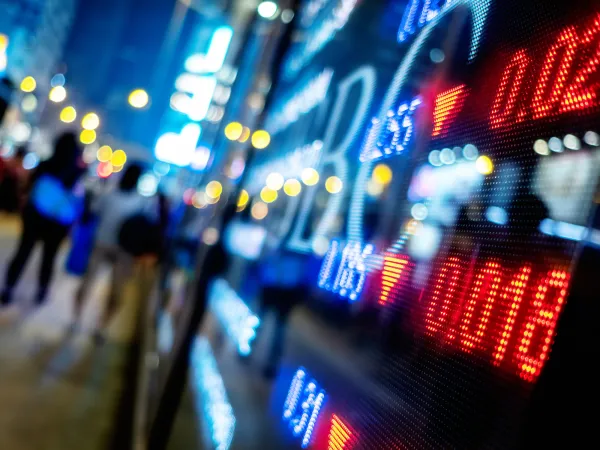 A digital stock market board displays red downward arrows and numbers at night with blurred city lights and pedestrians in the background.