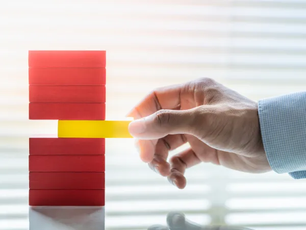 A hand in a blue sleeve pulls a yellow block from a stack of red wooden blocks, with blinds and sunlight in the background.