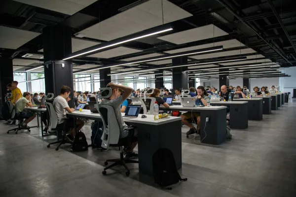 Modern open-plan office with rows of developers working on laptops at shared desks under sleek ceiling lighting.