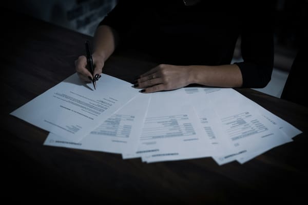 A person's hands signing financial documents spread across a dark desk