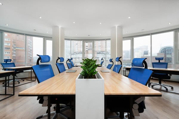 Empty open-plan office with a long shared desk, blue mesh chairs and floor-to-ceiling windows overlooking a city
